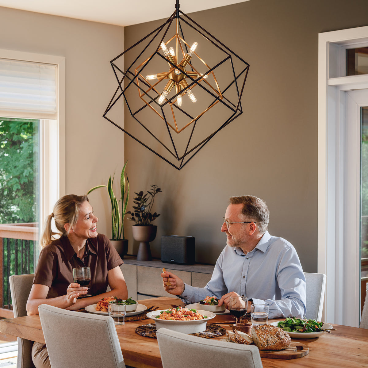 A couple dines under the Artika Imperium 9-light LED chandelier in a bright, modern dining room with plants and geometric decor.