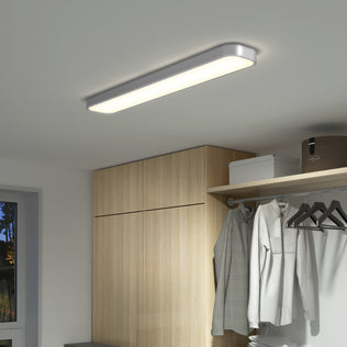 The Artika Europa 36 LED flush mount light with remote brightens a wardrobe area featuring wooden cabinets, hanging clothes, and a window that reveals the evening sky.