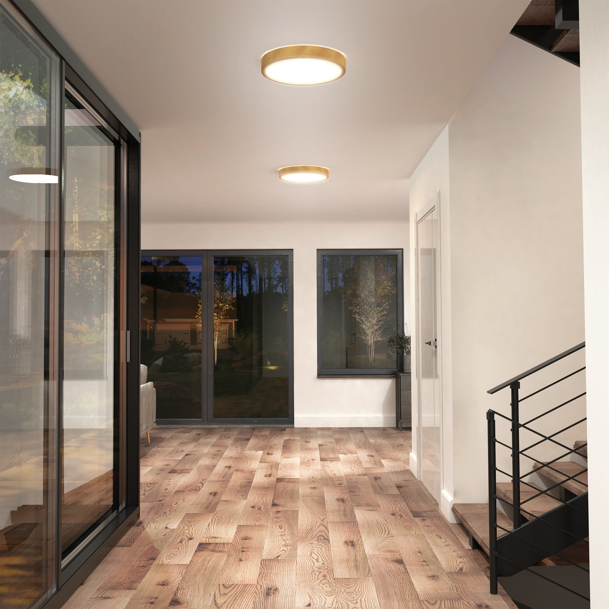 Modern hallway with wood floors, large glass windows and doors, black-framed staircase railing, and ceiling lights. Outside, trees and a garden are visible through the windows at dusk.