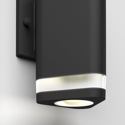 The Artika Trygon indoor-outdoor wall light features a modern black rectangular design with rounded edges and a frosted top section that emits soft white light, mounted on a plain light-colored wall.