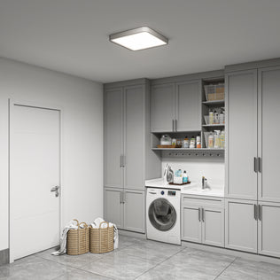 A modern laundry room features gray cabinets, a front-loading washer, sleek countertops with a sink, open shelves for cleaning supplies, wicker baskets holding towels on tiled floors, and is illuminated by the Artika Europa 18 in. LED flush mount light with remote.