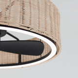 Close-up of a ceiling fan with black blades and a circular, wicker-patterned light fixture. The light glows around the edge of the woven shade, highlighting its texture against a light background.