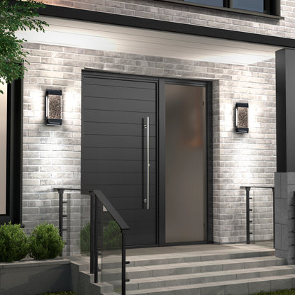 Modern front entrance with a large matte black door, vertical handle, frosted glass side panel, gray brick walls, steps with metal railings, and Artika Essence Glow Box indoor-outdoor LED wall lights on either side of the doorway.