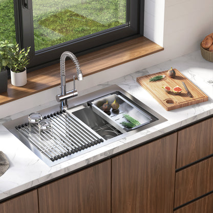 The Artika Newport double bowl stainless steel sink features a spring faucet, roll-up drying rack, and cutting board, set on a marble countertop with fresh produce, utensils, wooden cabinets, and a window overlooking greenery.
