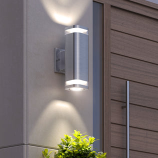 The Artika Mettle LED indoor-outdoor wall light, mounted on a light wall near a wooden door, casts white light beams up and down, with green plants visible below.