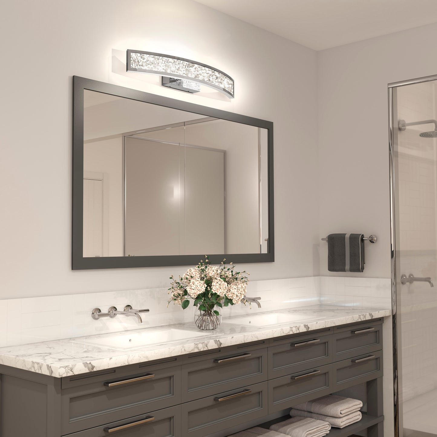 Modern bathroom featuring a large mirror, marble countertop, gray double-sink vanity, Artika Glitter LED bathroom vanity light, fresh flowers in a vase, neatly folded towels, and a glass shower door in the background.