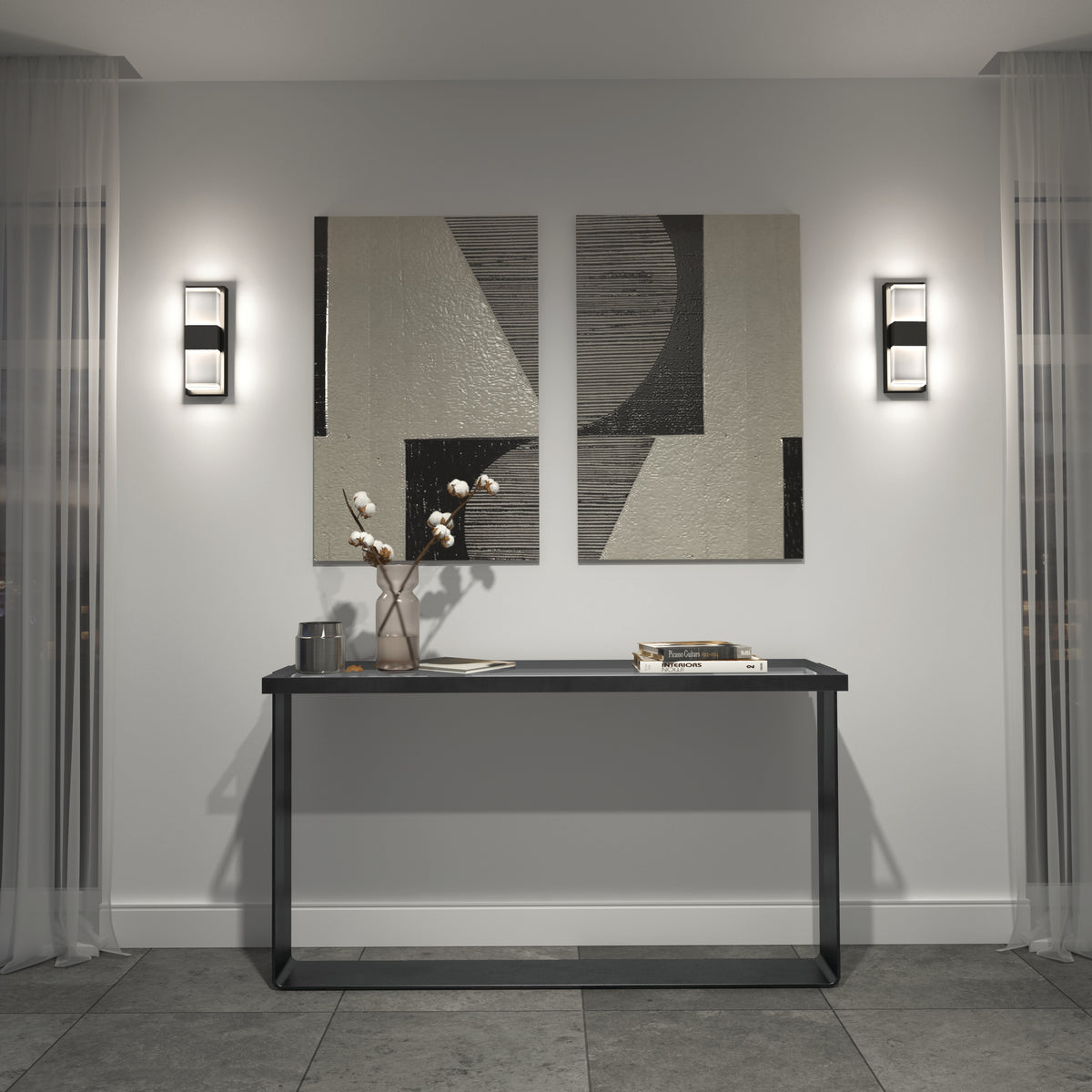 A modern entryway with a slim black console table holding a vase with cotton branches, books, and a small tray, set against a light wall with two abstract art pieces and two wall sconces, flanked by sheer curtains.