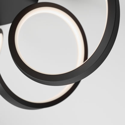 A close-up of the Artika Swirl Ribbon LED pendant light showcases two intertwined black metal rings with integrated LEDs, casting a soft white glow against a plain light background.