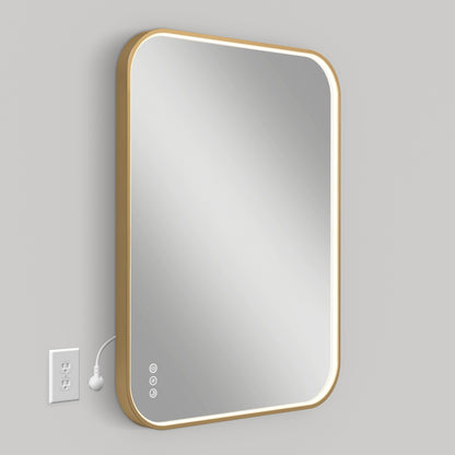 The Artika Royce Dimmable LED Mirror (3 CCT) is mounted on a light gray wall, plugged into a nearby outlet. Its rectangular shape features rounded corners, a gold frame, bottom-left touch controls, and adjustable color temperature.