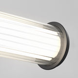 Close-up of a modern wall-mounted light fixture with a cylindrical, ribbed glass cover and a dark circular base, emitting bright white light against a neutral gray background.