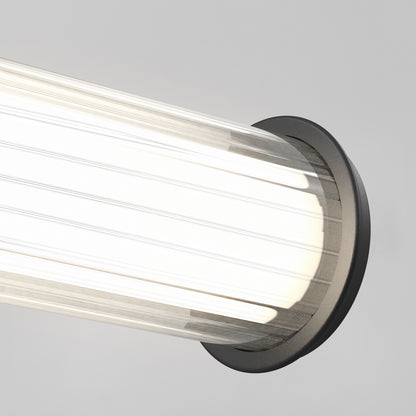 Close-up of the Artika Kendra LED contemporary bathroom vanity light 3 CCT, featuring a cylindrical ribbed glass shade and dark circular base, emitting bright white light against a plain light gray background.