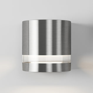 The Artika Valor Round LED indoor-outdoor wall light features a modern cylindrical brushed metal design with a horizontal frosted band emitting soft white light, shown mounted on a plain light gray wall.