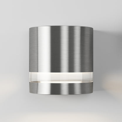 The Artika Valor Round LED indoor-outdoor wall light features a modern cylindrical brushed metal design with a horizontal frosted band emitting soft white light, shown mounted on a plain light gray wall.