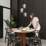 A woman arranges bowls and plates on a round wooden dining table with black chairs in a modern dining room featuring dark paneled walls, pendant lights, and a large potted plant.