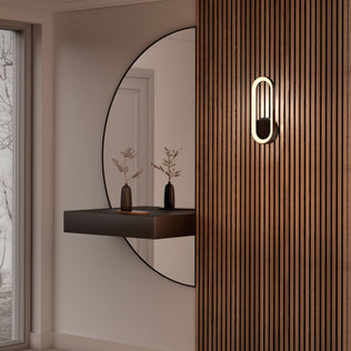 A minimalist interior features a large round mirror, a floating black shelf with vases and branches, and the Artika Aiden modern LED indoor-outdoor wall light (5 CCT) mounted on a vertical wooden slat wall. Natural light enters from a nearby window.