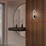 A modern entryway features a large round mirror, a floating black shelf with a small vase and dried branches, vertical wooden slats on the wall, and a sleek, oval wall sconce. A window reveals a wintery outdoor scene.