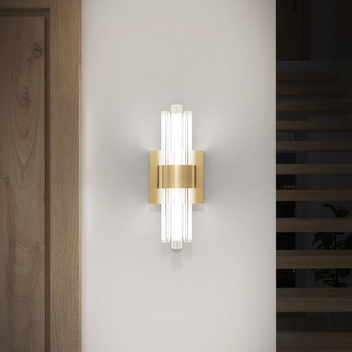 A modern wall sconce with a gold metallic center and vertical glass rods emits soft white light, mounted on a white wall near a wooden door and staircase.