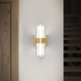 A modern wall sconce with a gold metallic center and vertical glass rods emits soft white light, mounted on a white wall near a wooden door and staircase.