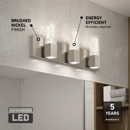 The Artika Hologram LED modern bathroom vanity light 5 CCT features three rectangular LED lights, a brushed nickel finish, energy efficiency labels, and a 5-year warranty. In the background are a mirror and shelves with towels.