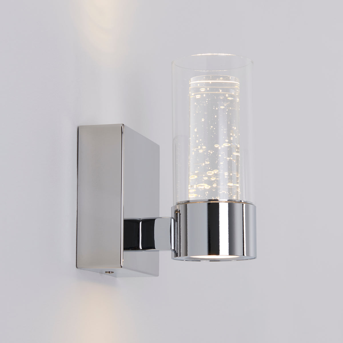 A modern wall sconce with a square chrome base and a cylindrical glass shade containing decorative bubbles, mounted on a white wall and illuminated.
