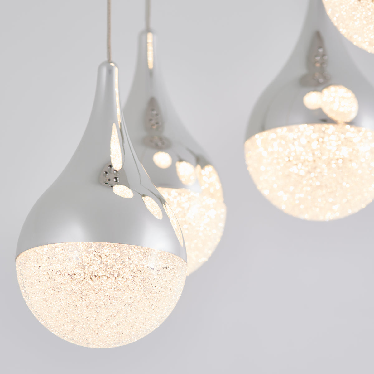 Close-up of modern pendant lights with shiny silver tops and sparkling, illuminated glass bottoms, hanging against a light gray background.