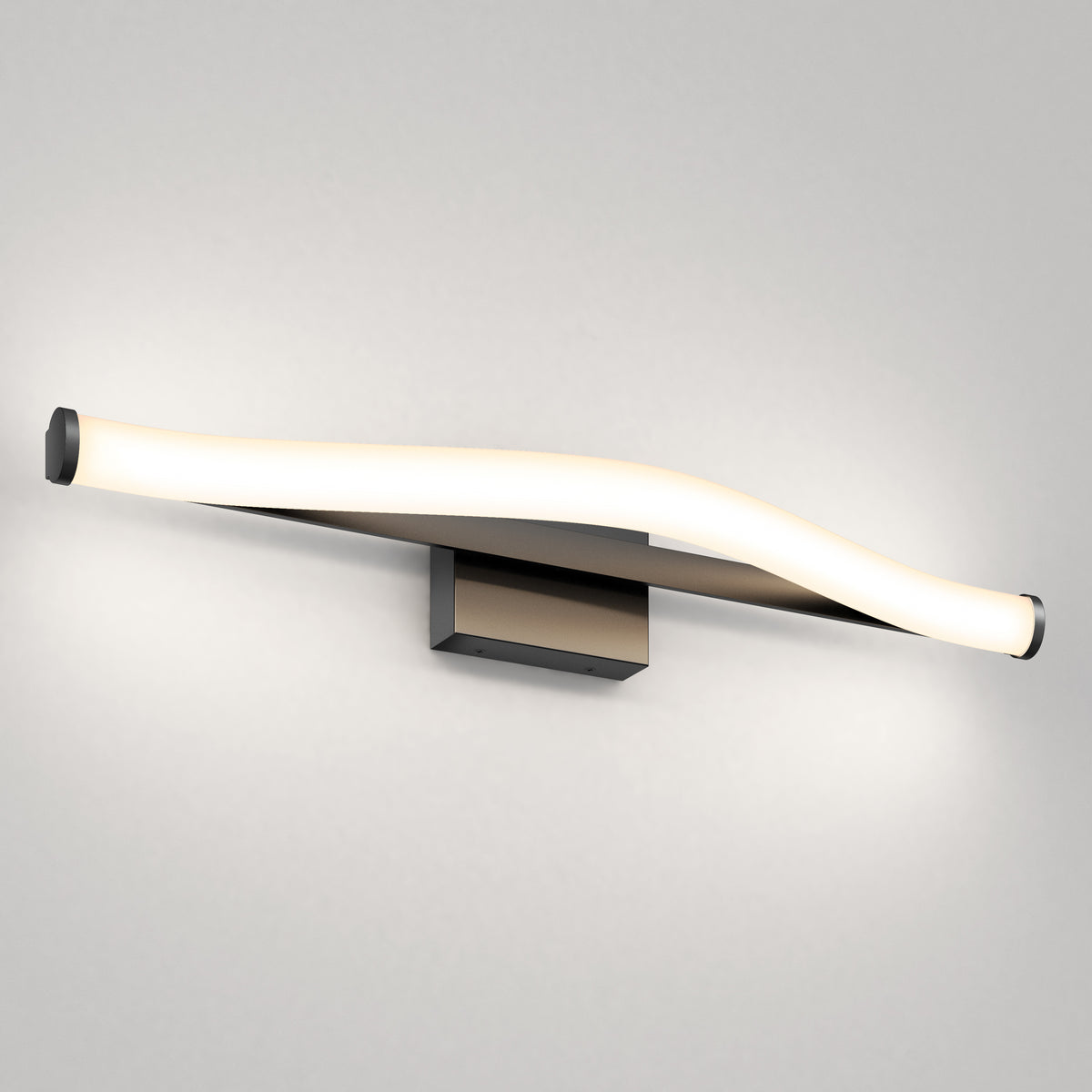A modern wall-mounted light fixture with a gently curved, elongated LED design, emitting a soft white glow against a plain light gray wall. The base is rectangular and metallic.