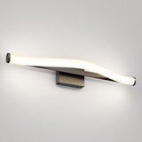 A modern wall-mounted light fixture with a gently curved, elongated LED design, emitting a soft white glow against a plain light gray wall. The base is rectangular and metallic.