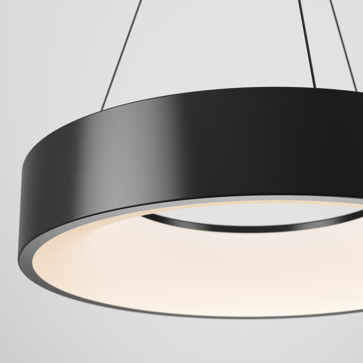 A close-up view of a modern, circular black pendant light fixture with a smooth finish, suspended by thin cables against a plain white background. The light glows softly from the inner edge.
