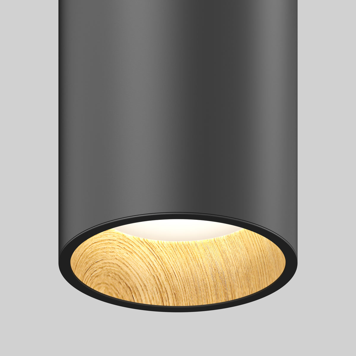 A close-up of a modern cylindrical ceiling light with a matte black exterior and a wooden textured inner rim, emitting a soft, warm white glow against a plain gray background.