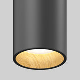 A close-up of a modern cylindrical ceiling light with a matte black exterior and a wooden textured inner rim, emitting a soft, warm white glow against a plain gray background.
