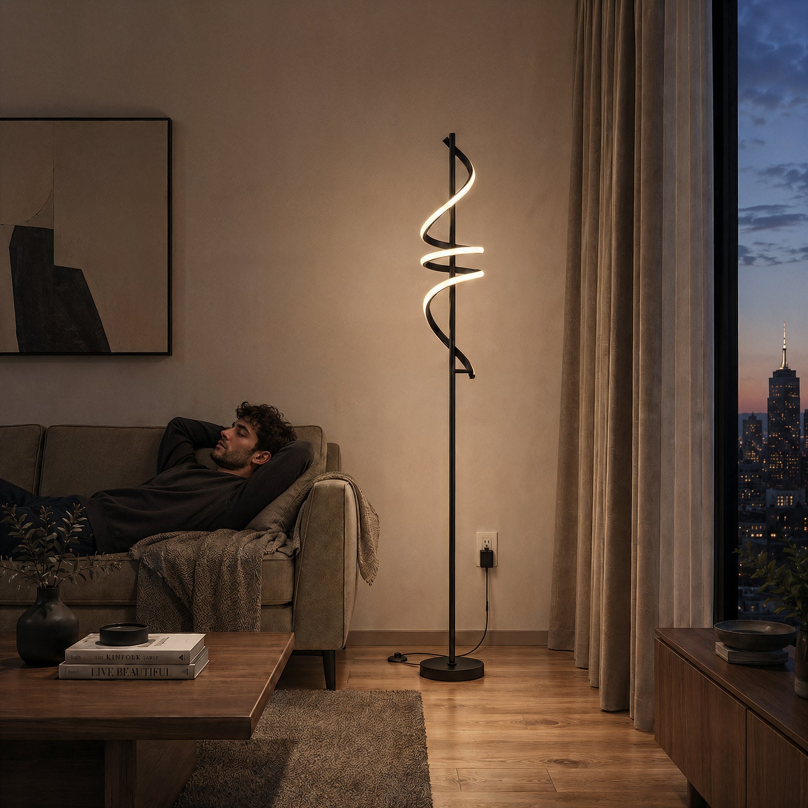 A man relaxes on a sofa in a modern living room at sunset. The Artika Coda modern LED floor lamp 5CCT glows beside him, while a large window reveals a city skyline with the Empire State Building in the background.