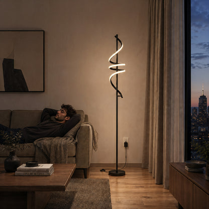 A man relaxes on a sofa in a modern living room at sunset. The Artika Coda modern LED floor lamp 5CCT glows beside him, while a large window reveals a city skyline with the Empire State Building in the background.