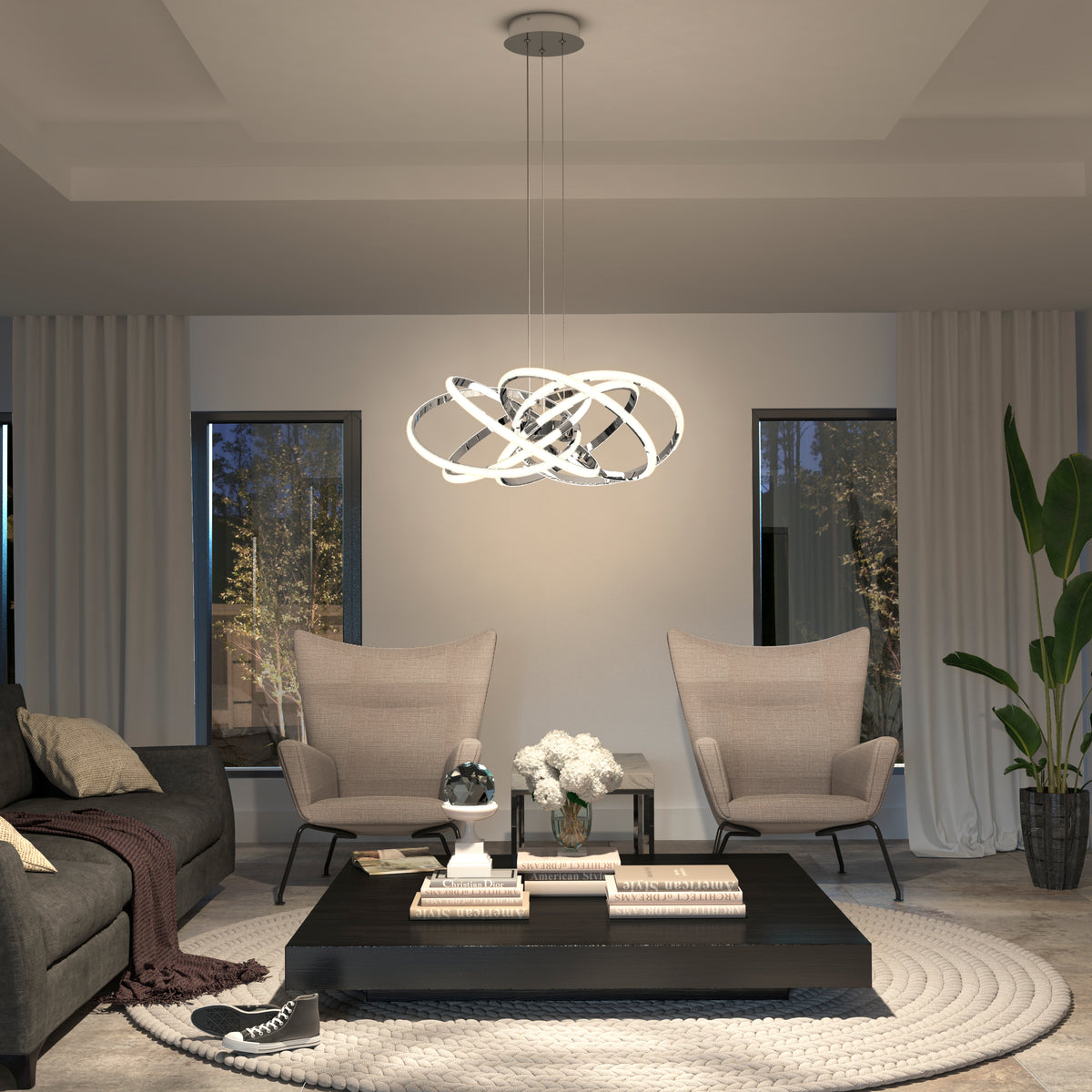 A modern living room with two beige armchairs, a dark sofa, a square black coffee table with books and flowers, a large round rug, and a sculptural pendant light hanging above. Two windows and a tall plant are in the background.