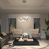 A modern living room with two beige armchairs, a dark sofa, a square black coffee table with books and flowers, a large round rug, and a sculptural pendant light hanging above. Two windows and a tall plant are in the background.