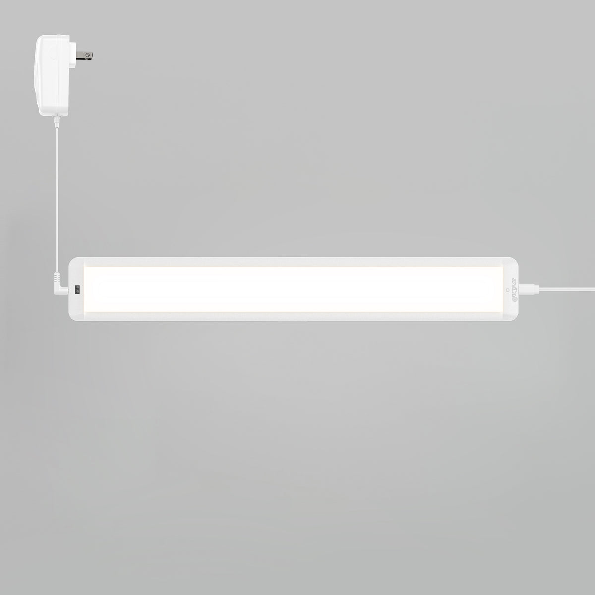 A rectangular white LED light fixture is mounted on a light gray wall, connected to a white power adapter with a cable. The light is turned on, emitting a bright glow.