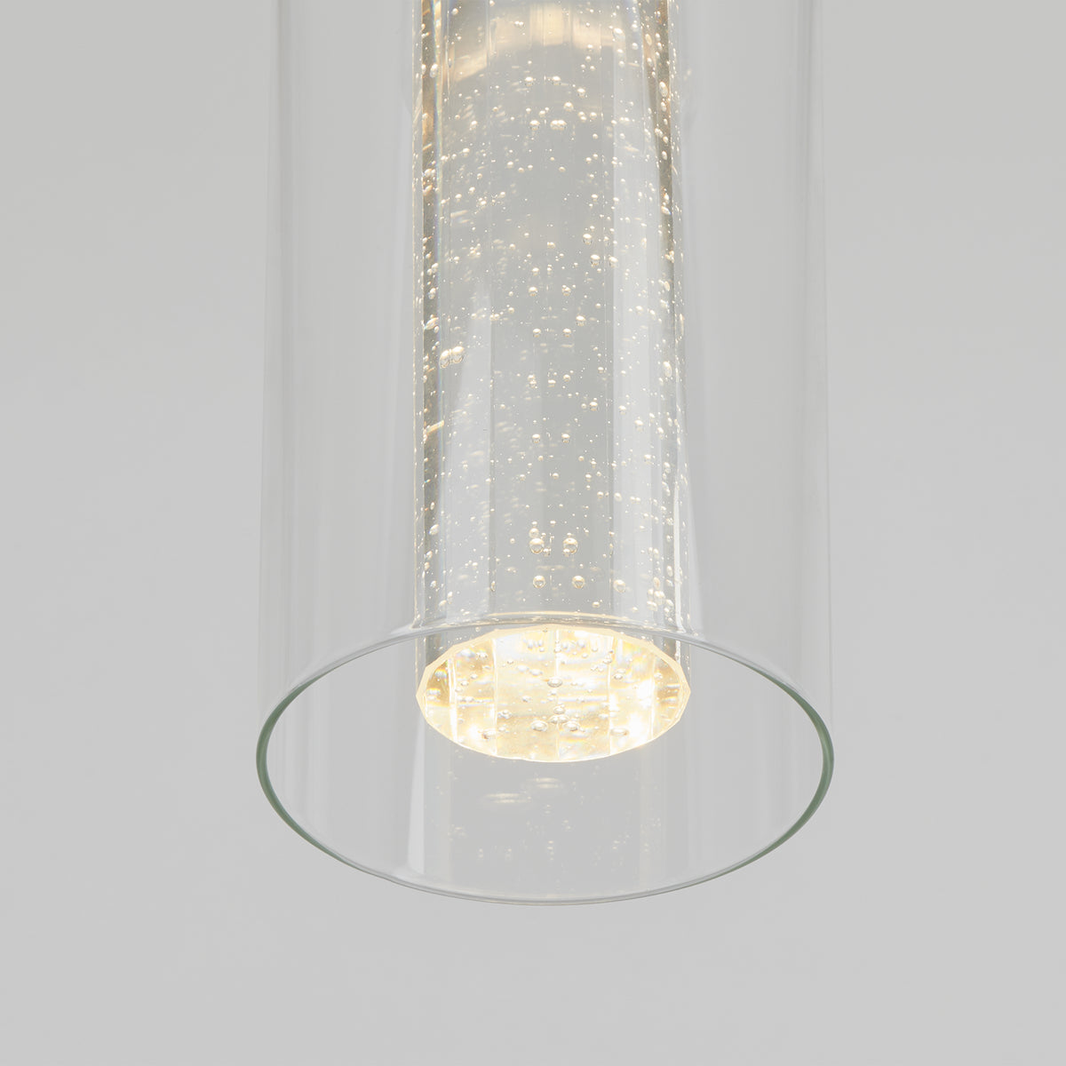 A close-up of a modern cylindrical glass pendant light with tiny bubbles inside, emitting a soft, warm glow against a plain light gray background.