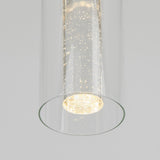 A close-up of a modern cylindrical glass pendant light with tiny bubbles inside, emitting a soft, warm glow against a plain light gray background.