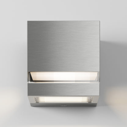 The Artika Valor Square LED indoor-outdoor wall light features a modern square design with brushed metal and a central illuminated strip, mounted on a white wall, casting a soft downward and outward glow.