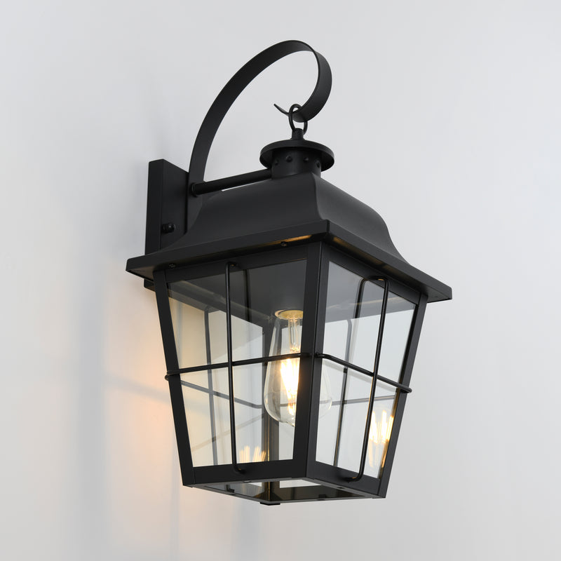 The Artika Jatoba outdoor lantern wall light features a black metal frame, curved top handle, and clear glass panels, showcasing a glowing bulb inside and is shown mounted on a white wall.