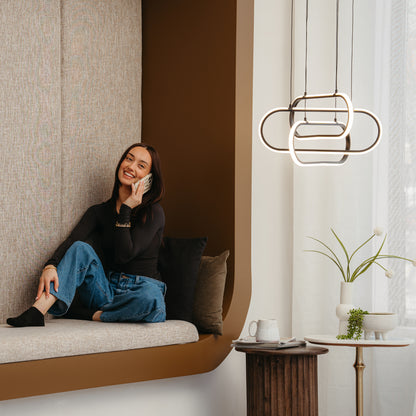 A woman smiles and talks on her phone, seated on a cushioned bench in a cozy room. Beside her, an Artika Lewis modern LED pendant light (3 CCT) hangs above a small table with a plant and cup, adding style to the space.