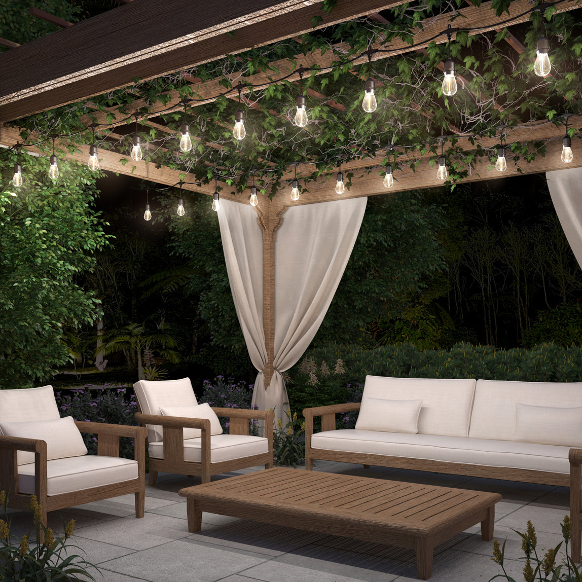 A cozy outdoor patio at night features wooden furniture with cream cushions, a pergola draped in Artikas Blaze 20 LED Outdoor Solar String Lights and ivy, sheer curtains, lush greenery, and a central wooden coffee table.