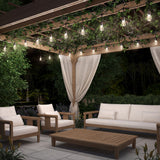 A cozy outdoor patio at night with string lights, a wooden pergola draped with ivy, cream-colored cushioned chairs, a sofa, a wooden coffee table, and white curtains, surrounded by greenery.