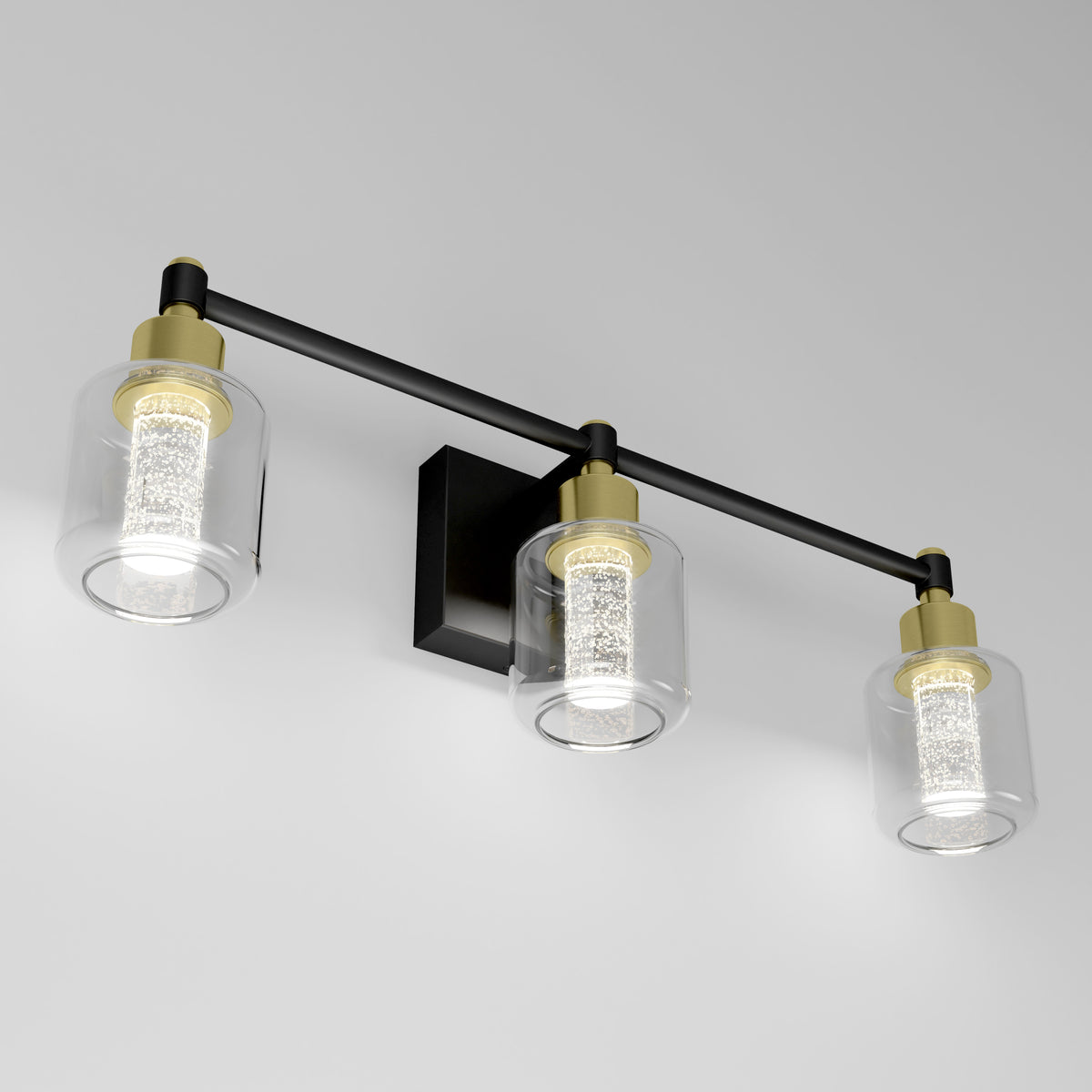Three modern wall-mounted light fixtures with cylindrical glass shades and gold accents are attached to a black bar on a white wall. Each light has a textured inner cylinder that diffuses the glow.