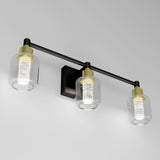 Three modern wall-mounted light fixtures with cylindrical glass shades and gold accents are attached to a black bar on a white wall. Each light has a textured inner cylinder that diffuses the glow.