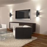 Modern living room with a wall-mounted TV, two black and white armchairs, a glass coffee table, a white rug, wooden floors, a dark cabinet, and two wall sconces with soft lighting on either side of the TV.