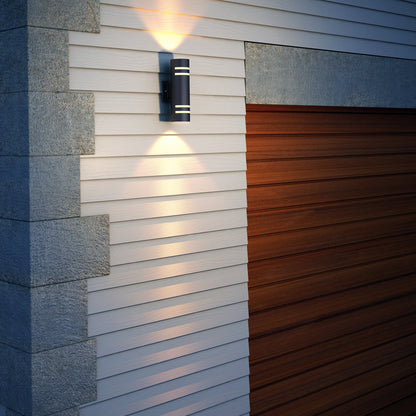 The Artika V3 indoor-outdoor wall light is mounted on white siding near a wooden garage door, casting warm up and down illumination on the wall.