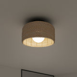 A round ceiling light with a woven, beige lampshade emits a soft glow, mounted on a smooth white ceiling above a partially visible gray-framed picture.