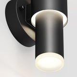 Close-up of a modern black cylindrical wall sconce with a round base, emitting a soft white light from the bottom. The fixture has a sleek, minimalist design against a light background.