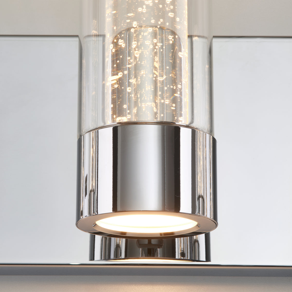 A close-up of a modern light fixture with a polished chrome base and a cylindrical glass shade featuring small bubbles inside, mounted on a reflective surface. The light inside is turned on.