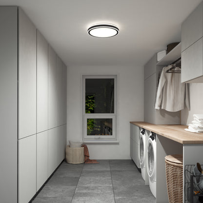 A modern, minimalist laundry room features gray cabinets, a wooden countertop, washer and dryer, laundry basket, hanging clothes, and a window with greenery. The Artika Altitude 15 in. LED flush mount light brightens the space.
